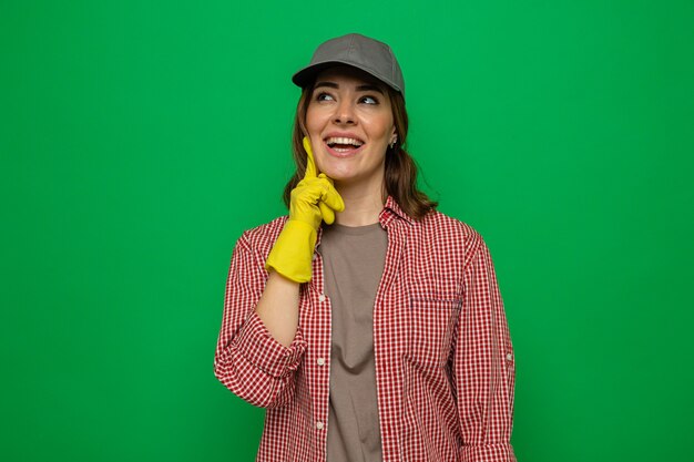 緑の背景の上に立っていることを夢見て元気に笑顔を見上げてゴム手袋を着用した格子縞のシャツとキャップの若い掃除の女性