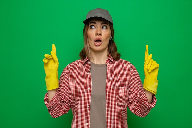 Young cleaning woman in plaid shirt and cap wearing rubber gloves looking up surprised pointin with index fingers up