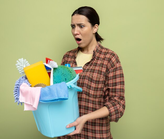 Young cleaning woman in plaid shirt holding bucket with cleaning tools looking aside amazed and shocked standing on green