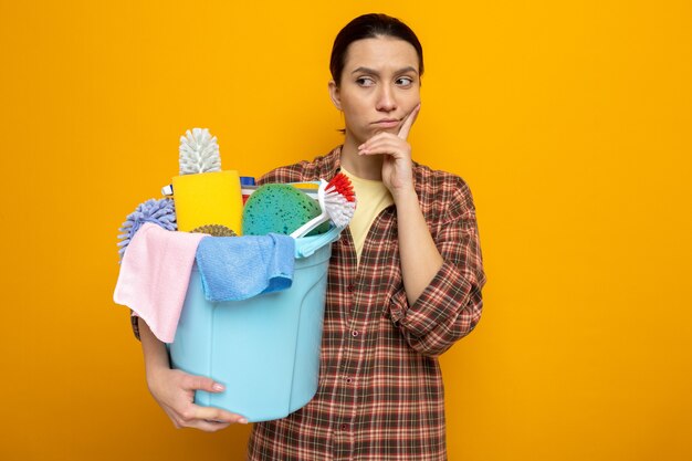 Young cleaning woman in plaid shirt holding bucket with cleaning tools looking aside with pensive expression on face thinking standing on orange