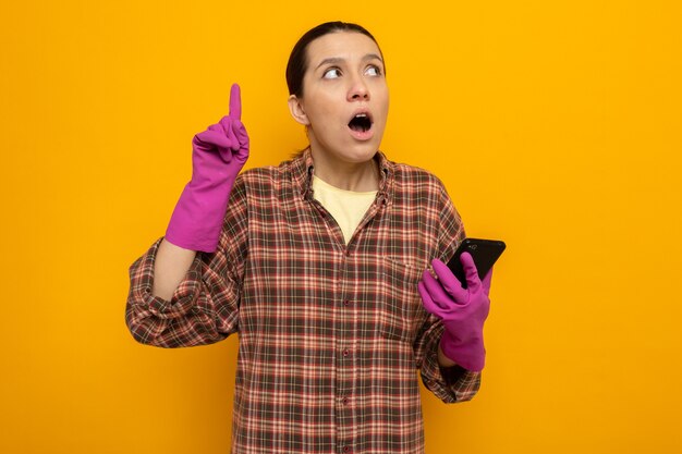 Молодая уборщица в клетчатой рубашке в резиновых перчатках, держащая смартфон, удивленно смотрит вверх, показывая указательный палец с новой идеей