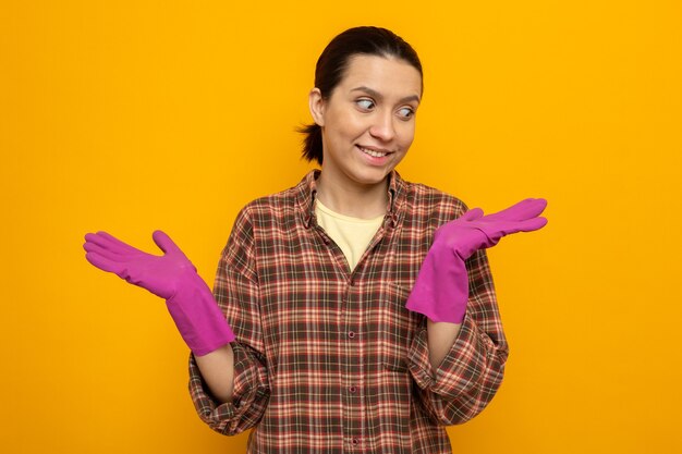 Молодая уборщица в клетчатой рубашке в резиновых перчатках смотрит в сторону, счастливая и позитивная, поднимая руки, весело улыбаясь