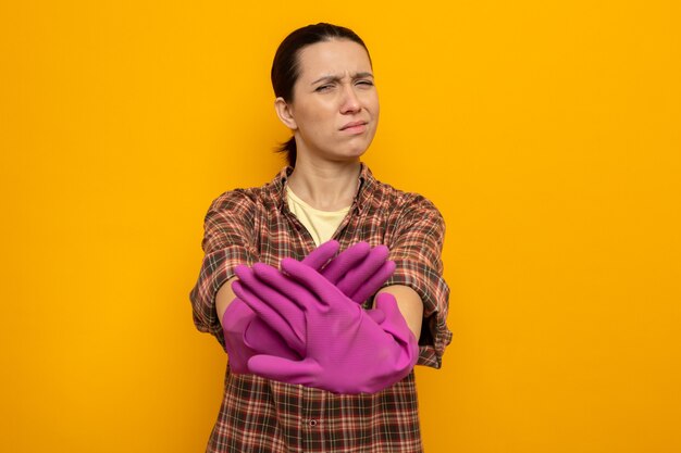 Young cleaning woman in plaid shirt in rubber gloves looking at front displeased making stop gesture with hands standing over orange wall