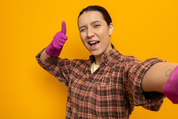 Young cleaning woman in plaid shirt in rubber gloves looking at front happy and positive smiling cheerfully showing thumbs up standing over orange wall