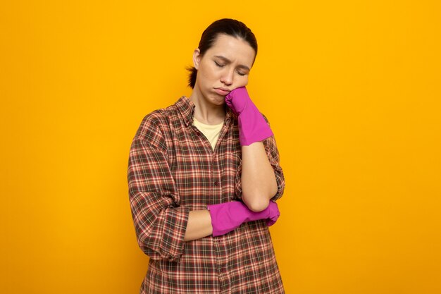 Young cleaning woman in plaid shirt in rubber gloves looking tired and bored leaning head on her fist standing on orange