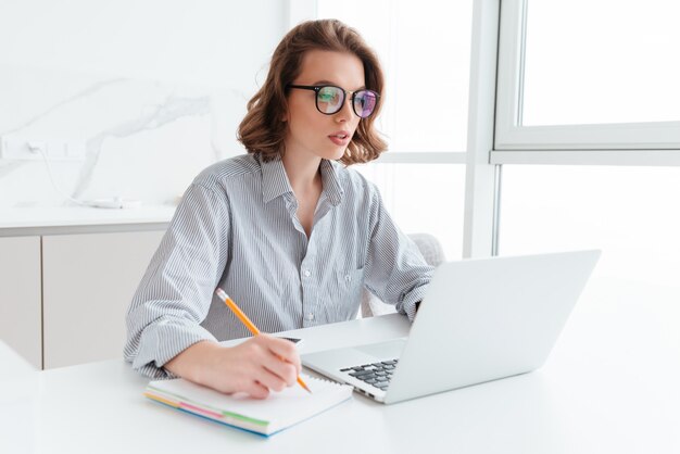 Young concentrated brunette woman in glasses wokking with laptop while sitting at table in light kitchen