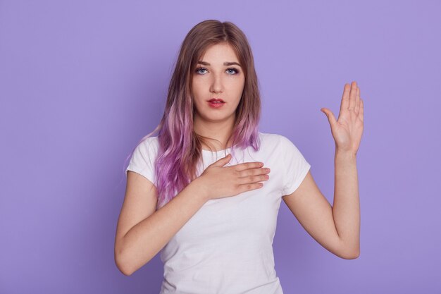 Young concentrated female looking directly at camera, keeping one hand on chest and raising other one, showing palm, swearing, isolated over purple wall.