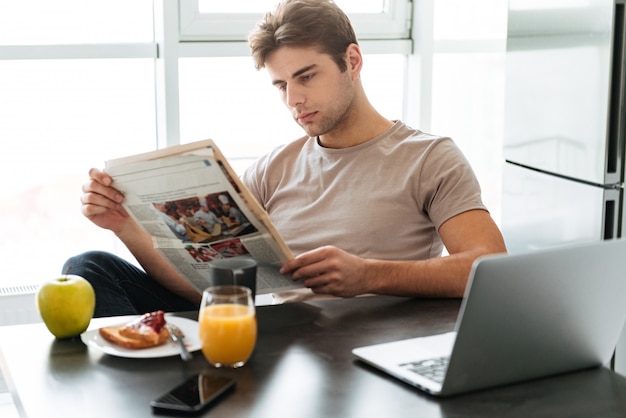 Young concentrated man reading newspaper while sitting in kitchen