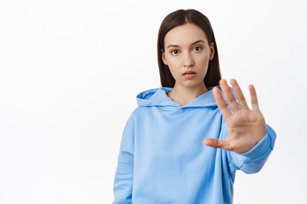 Young concerned woman extend hand and tell no, disagree, forbid or reject smth, taboo gesture, standing in hoodie against white wall.