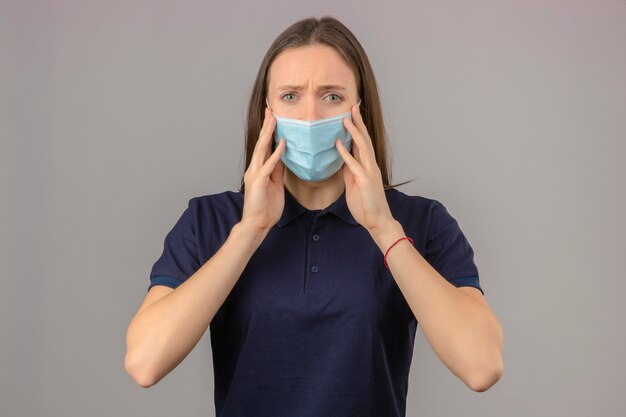 Young  concerned and worried woman wearing blue t shirt  in protective medical mask looking at camera with raised hands touching her mask on light grey isolated background