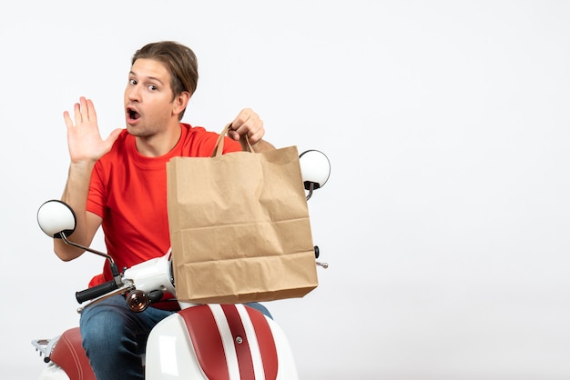 Young concerning courier guy in red uniform sitting on scooter holding paper bag on white wall