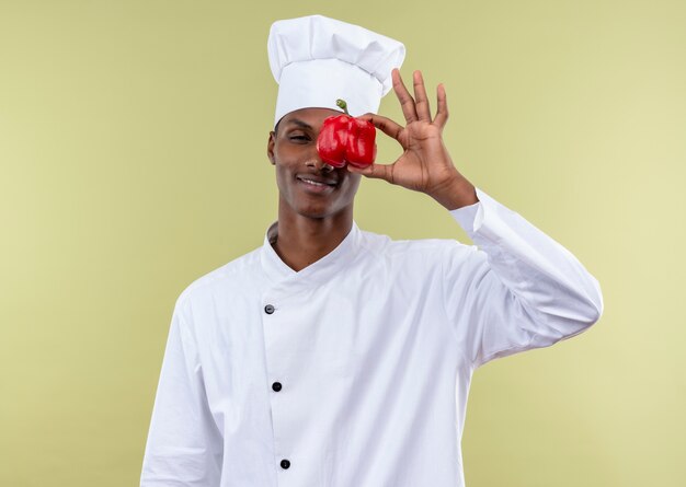 Молодой уверенный в себе афро-американский повар в униформе шеф-повара закрывает глаза красным перцем, изолированным на зеленом фоне с копией пространства