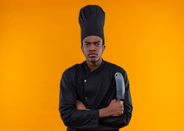 Young confident afro-american cook in chef uniform crosses arms and holds knife isolated on orange background with copy space