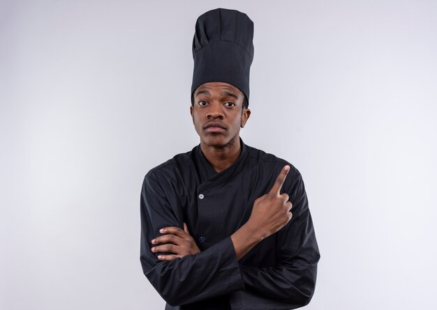 Молодой уверенный в себе афро-американский повар в униформе шеф-повара скрещивает руки и указывает вверх на белом фоне с копией пространства