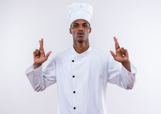 Young confident afro-american cook in chef uniform crosses fingers of both hands isolated on white background with copy space