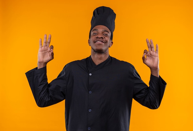 Молодой уверенный в себе афро-американский повар в униформе шеф-повара жестами показывает знак рукой обеими руками, изолированными на оранжевом фоне с копией пространства