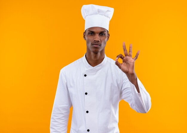 コピースペースでオレンジ色の背景に分離されたシェフの制服のジェスチャーで若い自信を持ってアフリカ系アメリカ人の料理人