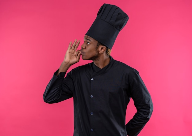 Young confident afro-american cook in chef uniform gestures tasty delicious isolated on pink background with copy space