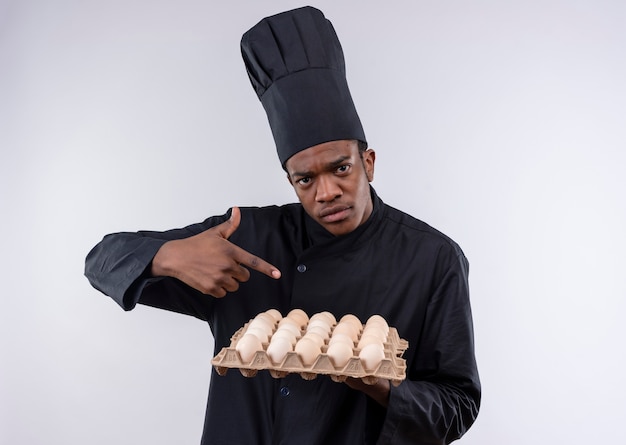 Young confident afro-american cook in chef uniform holds batch of eggs and points with finger isolated on white background with copy space