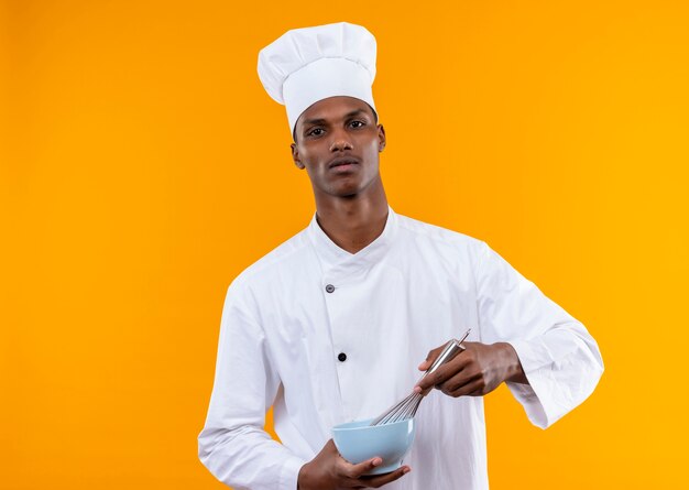 Young confident afro-american cook in chef uniform holds bowl and whisk isolated on orange background with copy space