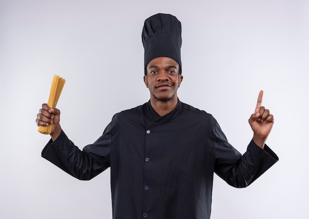 Young confident afro-american cook in chef uniform holds bunch of spaghetti and points up isolated on white background with copy space