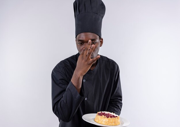 Il giovane cuoco afroamericano sicuro in uniforme del cuoco unico tiene la torta sulla zolla e sui gesti delizioso segno su bianco con lo spazio della copia