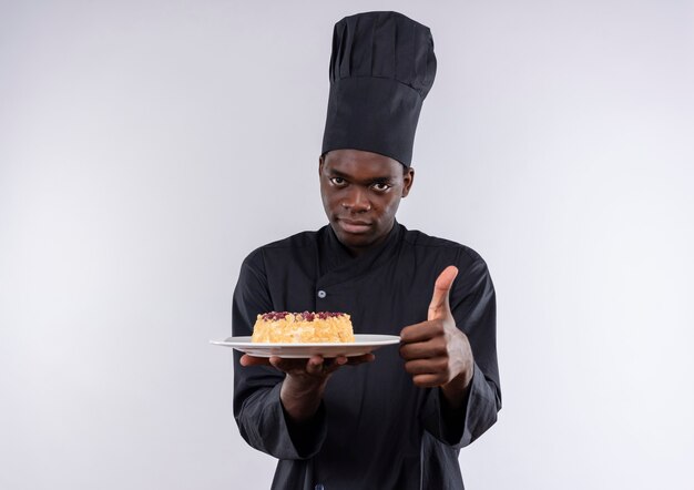 Young confident afro-american cook in chef uniform holds cake on plate and thumbs up on white  with copy space