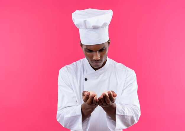 Young confident afro-american cook in chef uniform holds eggs in both hands isolated on pink background with copy space