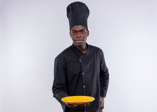 Young confident afro-american cook in chef uniform holds empty plate and puts hand on waist on white  with copy space