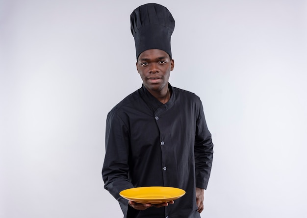 Молодой уверенный в себе афро-американский повар в униформе шеф-повара держит пустую тарелку и кладет руку на талию на белом с копией пространства