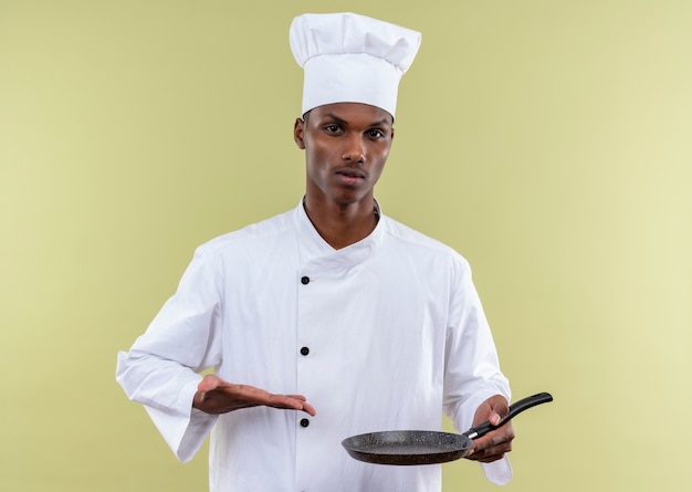 Young confident afro-american cook in chef uniform holds frying pan and points with hand isolated on green background with copy space