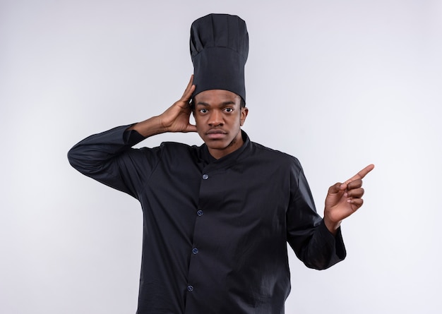 Young confident afro-american cook in chef uniform holds head and points to the side isolated on white background with copy space