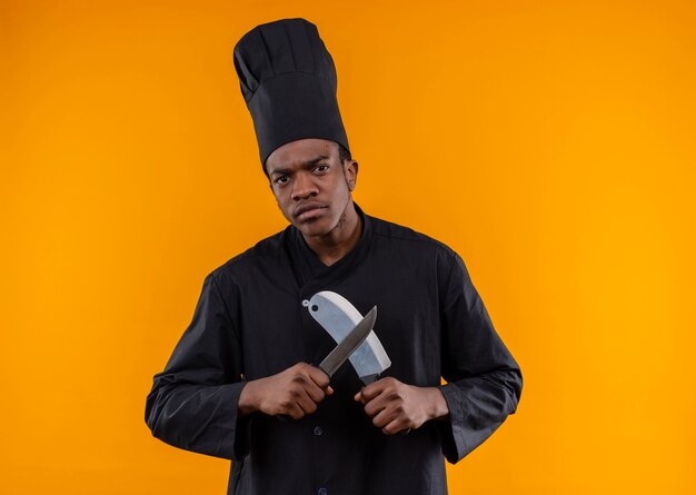 Young confident afro-american cook in chef uniform holds knifes isolated on orange background with copy space