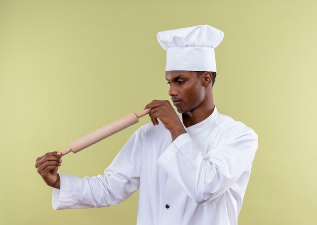 Young confident afro-american cook in chef uniform holds and looks at rolling pin isolated on green background with copy space