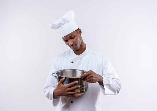Young confident afro-american cook in chef uniform holds and looks at saucepan on isolated white background with copy space