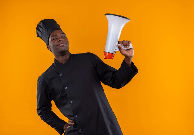 Young confident afro-american cook in chef uniform holds loud speaker on orange  with copy space