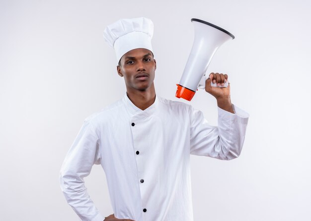 Young confident afro-american cook in chef uniform holds loud speaker and puts hand on waist isolated on white background with copy space