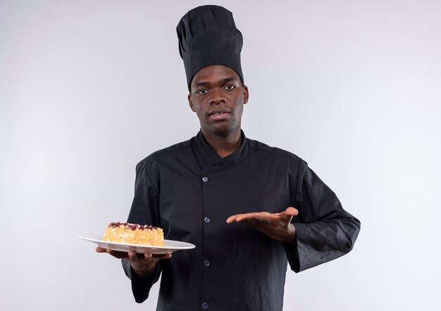 Young confident afro-american cook in chef uniform holds and points at cake on plate on white  with copy space