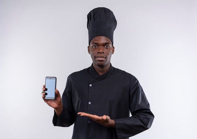 Young confident afro-american cook in chef uniform holds and points at phone on white  with copy space