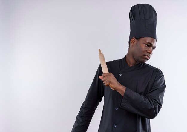 Молодой уверенный в себе афро-американский повар в униформе шеф-повара держит скалку и смотрит в сторону на белом с копией пространства