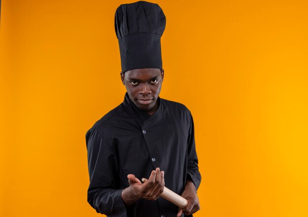 Young confident afro-american cook in chef uniform holds rolling pin on orange  with copy space