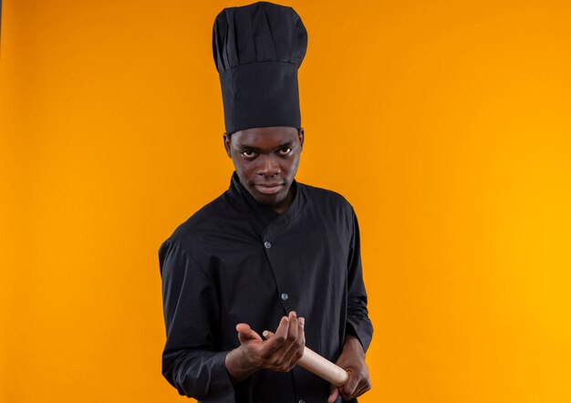 シェフの制服を着た若い自信のあるアフリカ系アメリカ人の料理人は、コピースペースでオレンジ色の麺棒を保持します