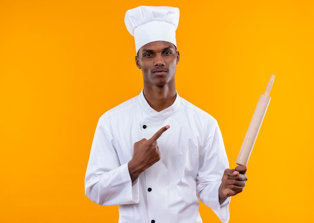 Young confident afro-american cook in chef uniform holds rolling pin and points at pin isolated on orange background with copy space