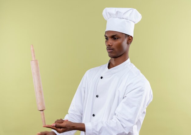 Young confident afro-american cook in chef uniform holds rolling pin straight on finger isolated on green background with copy space