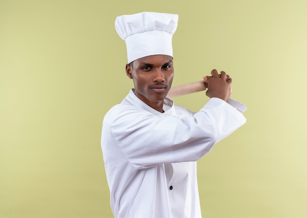 Young confident afro-american cook in chef uniform holds rolling pin behind with both hands isolated on green background with copy space