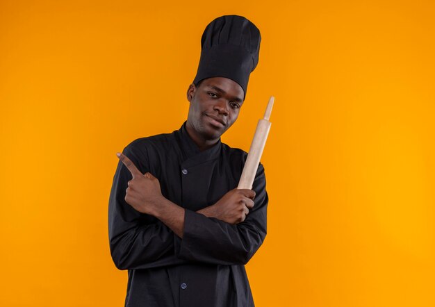 요리사 제복을 입은 젊은 자신감 아프리카 계 미국인 요리사는 복사 공간이 오렌지에 교차 팔을 가진 롤링 핀을 보유하고 있습니다.