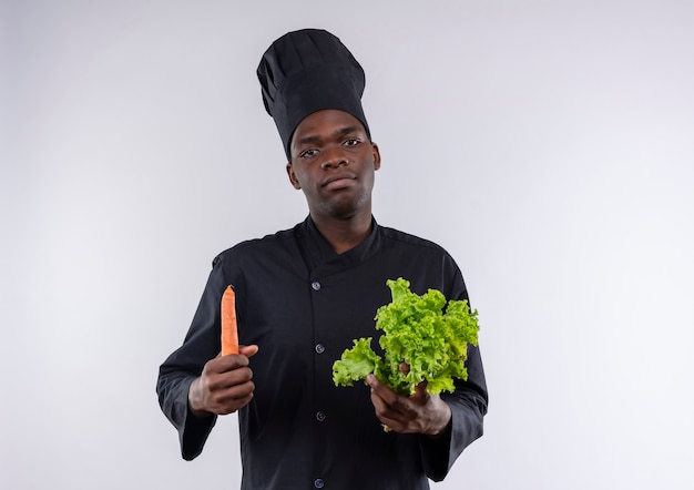 Young confident afro-american cook in chef uniform holds salad and carrot on white  with copy space