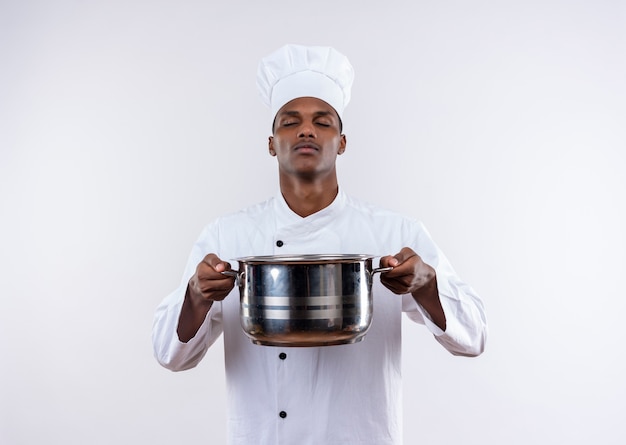 Young confident afro-american cook in chef uniform holds saucepan with closed eyes on isolated white background with copy space