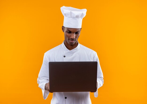 シェフの制服を着た若い自信を持ってアフリカ系アメリカ人の料理人は、コピースペースでオレンジ色の背景に分離されたラップトップを見て