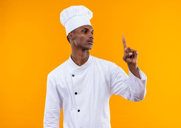 Young confident afro-american cook in chef uniform points up and looks at finger isolated on orange background with copy space